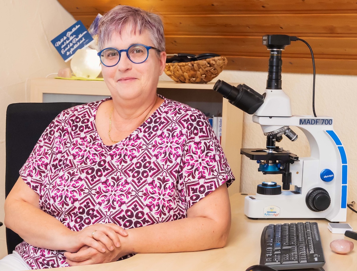 Heilpraktikerin Angelika Maria Zimmermann an ihrem Schreibtisch in ihrer Praxis in Ettringen. Sie lächelt freundlich und schafft eine einladende Atmosphäre für ein Patientengespräch.